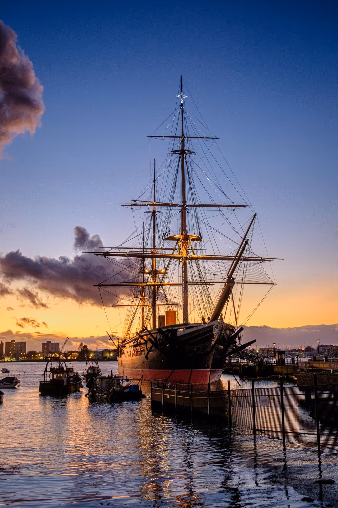 Historic large sailing ship docked at a harbor during sunset with city buildings in the background.