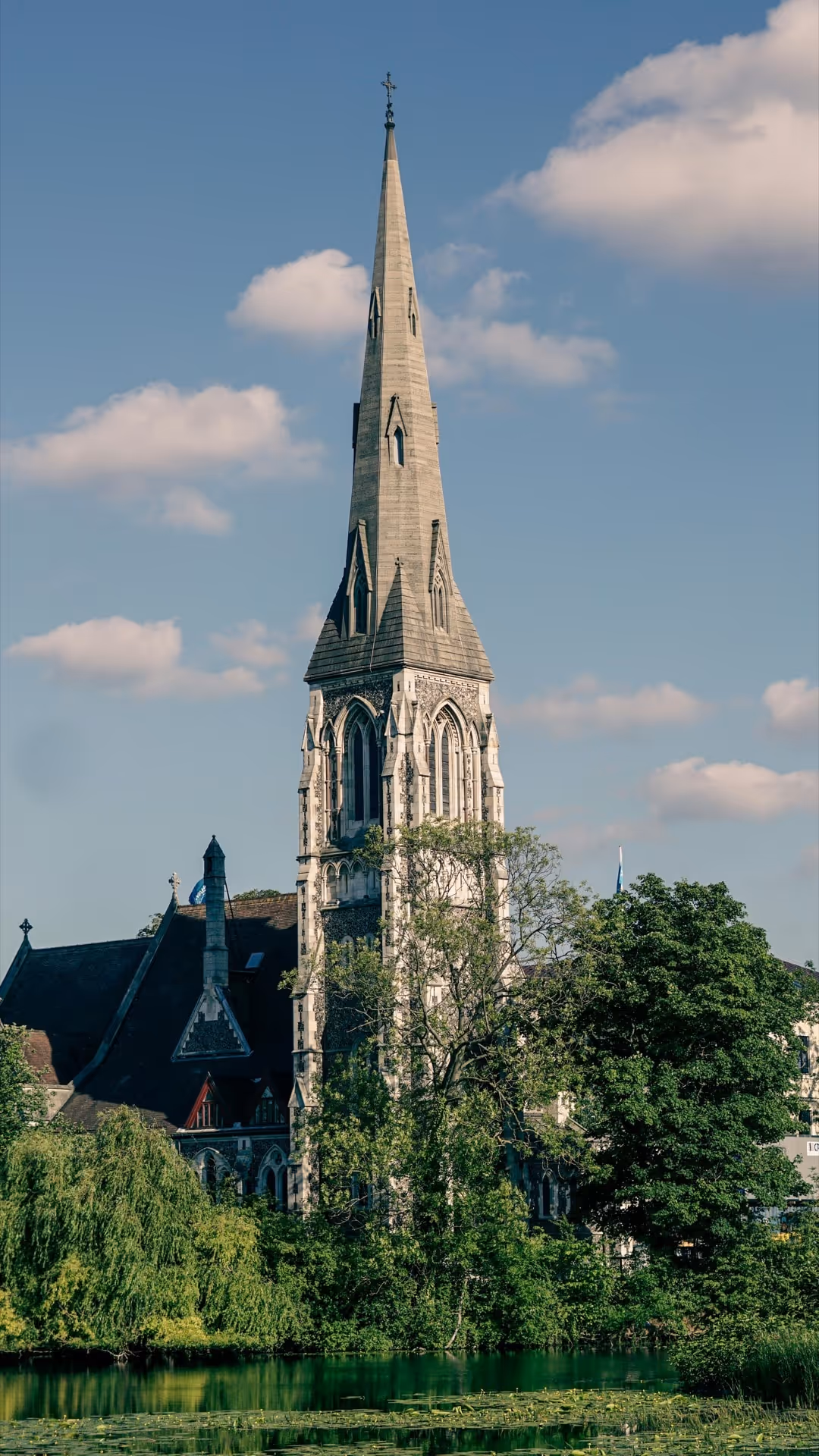 Tall, ornate church steeple surrounded by green trees beside a pond with lily pads under a partly cloudy sky.
