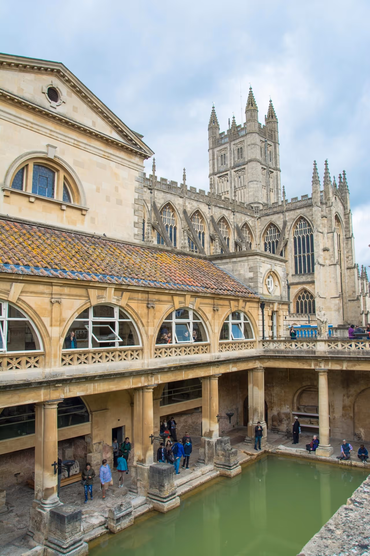 Visitors at the Roman Baths with the Gothic Bath Abbey in the background under a cloudy sky.
