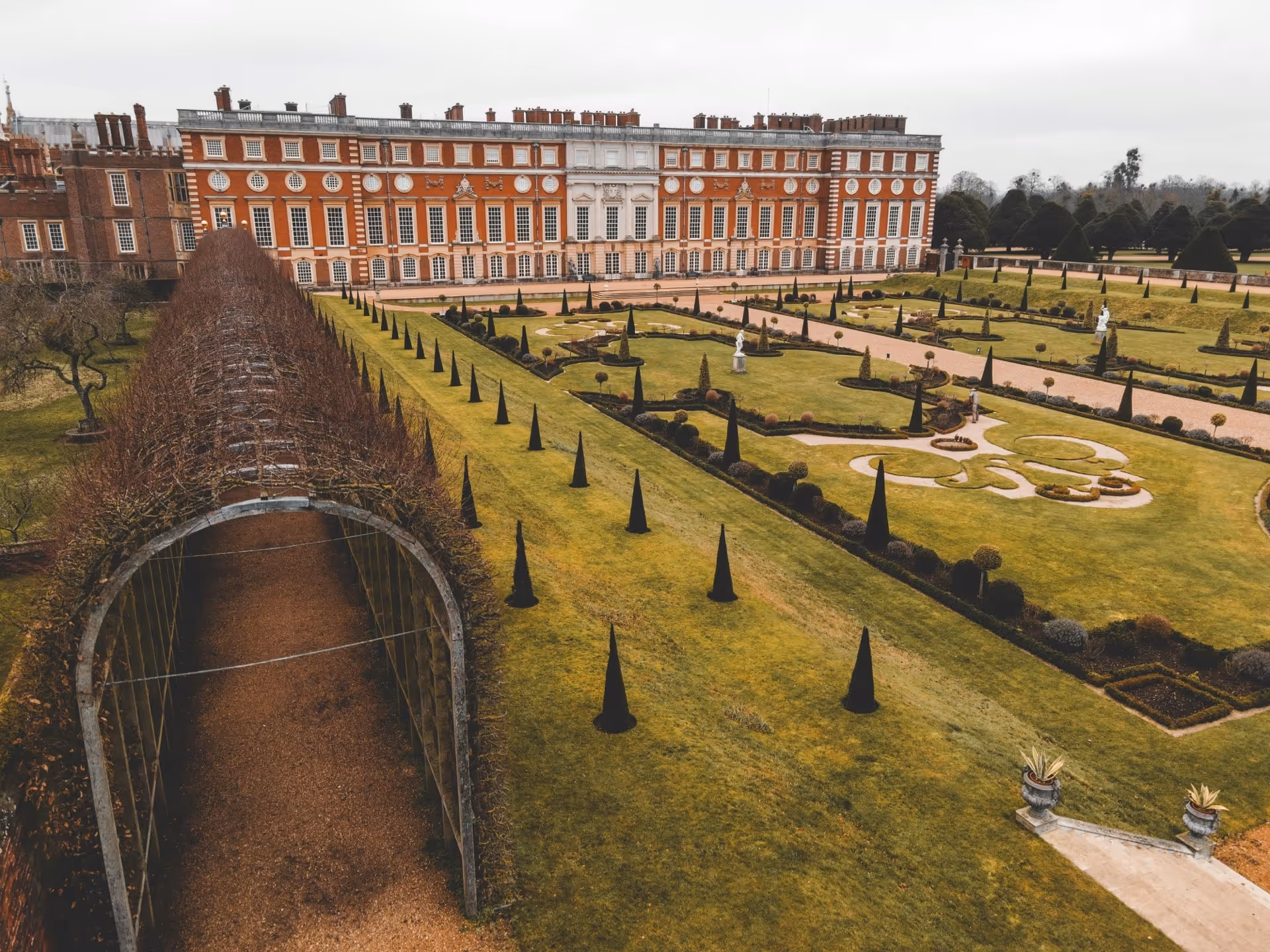 Formal garden with manicured lawns, pathways, conical topiary trees, classical statues, and a large historic brick and stone building in the background.
