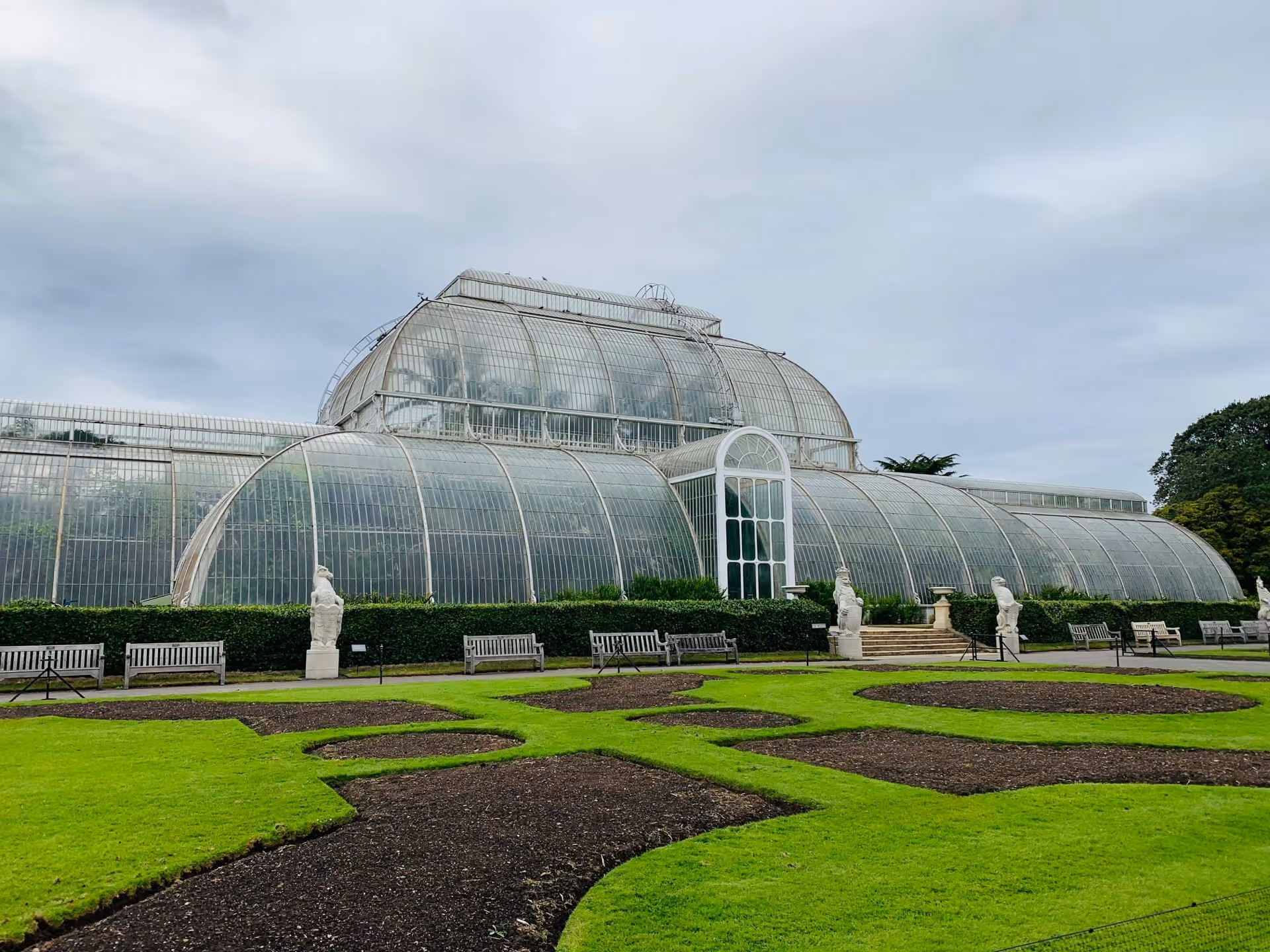 Large Victorian-style glass greenhouse with curved roof surrounded by manicured garden beds, benches, and stone statues under an overcast sky.