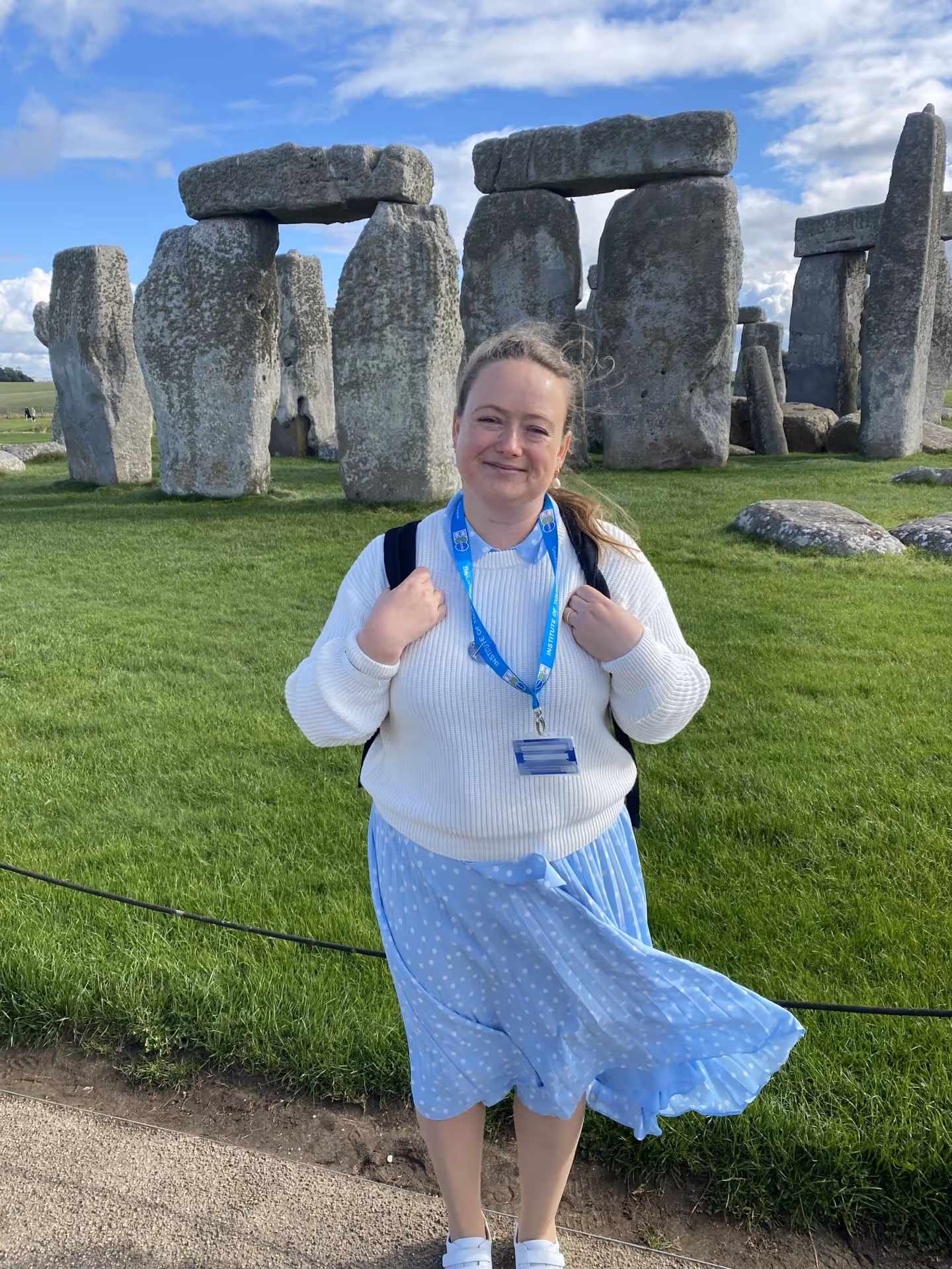 Woman wearing a white sweater and blue polka dot skirt with a lanyard, standing in front of Stonehenge under a partly cloudy sky.