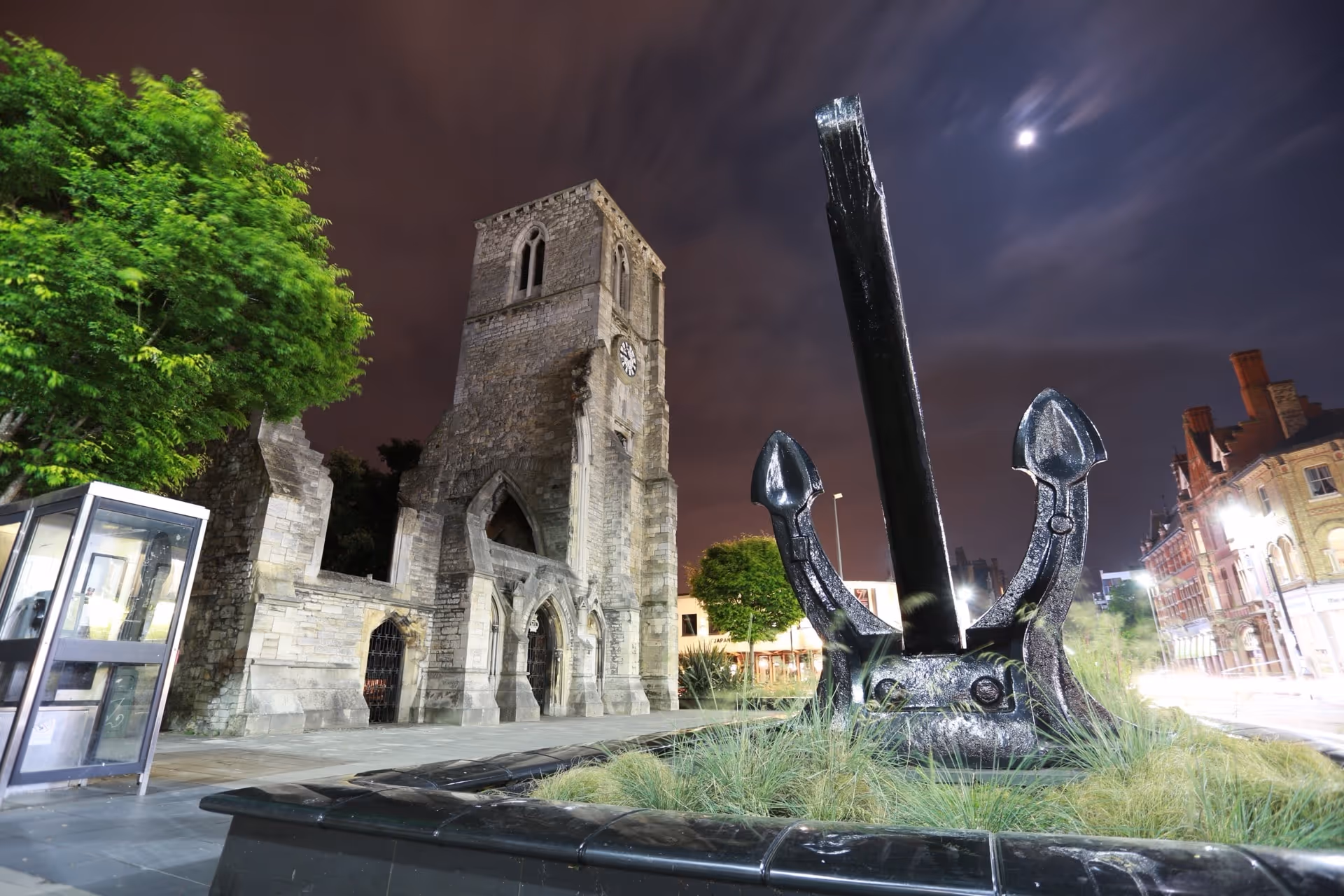 Night view of an old stone clock tower next to a large black anchor sculpture surrounded by grass in an urban setting.