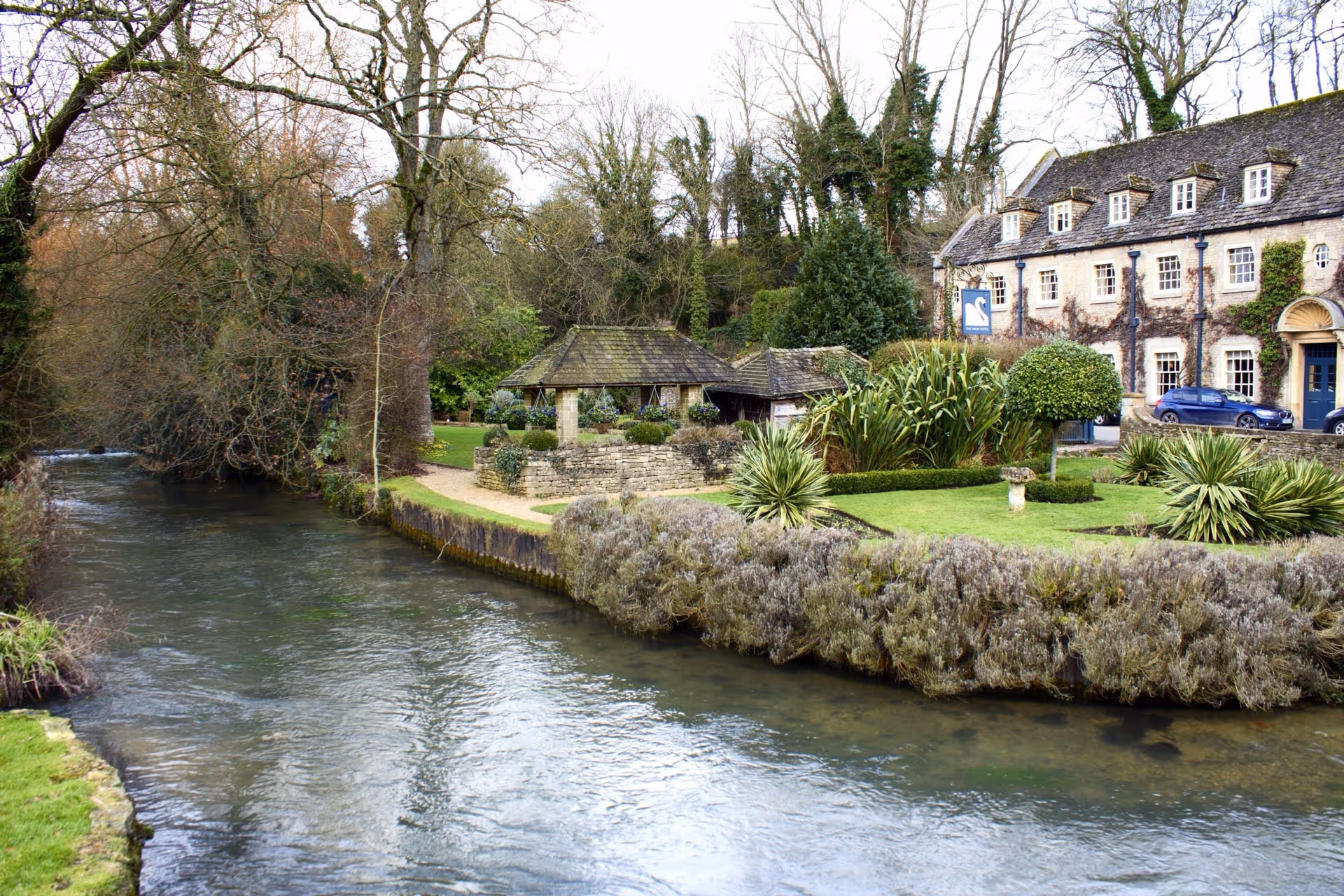 A peaceful stone building and garden with trimmed shrubs and trees next to a gently flowing river.