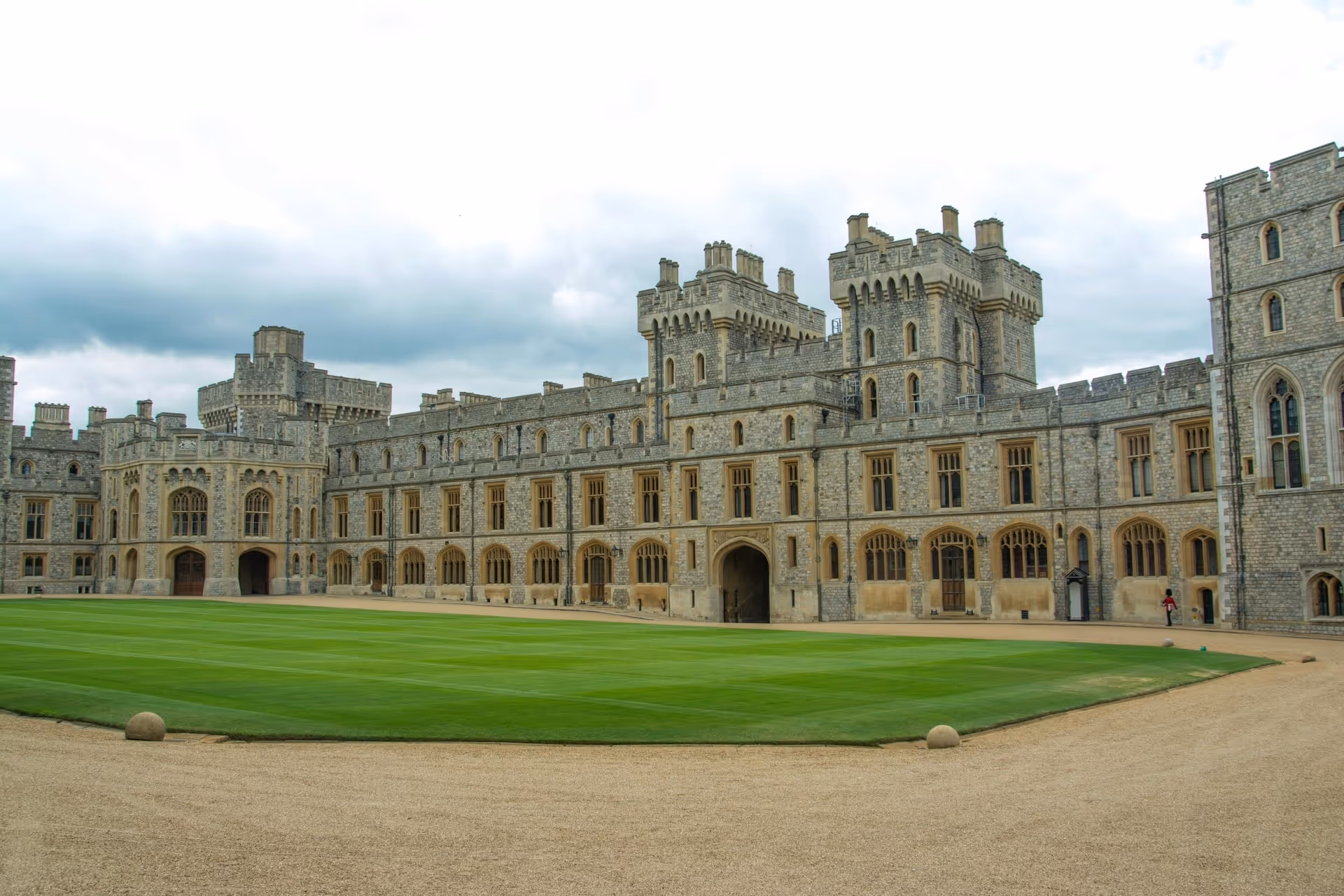 Large historic stone castle with tall towers overlooking a neatly manicured green lawn and gravel pathway under a cloudy sky.